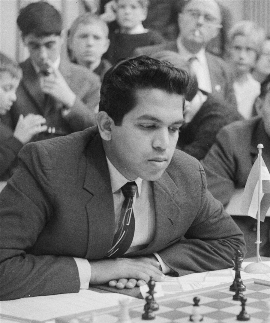 Black-and-white photograph of Manuel Aaron, a young man in a dark suit, leaning over a chessboard in concentration while spectators watch behind him.